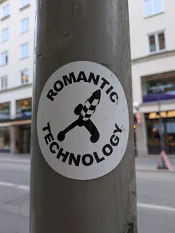 Street sticker A circular white sticker featuring a bold black illustration of a stylized rocket or phallic object in the center. The object has a checkered pattern on its tip and two small spherical elements at its base, resembling a joystick or a dildo. Curved around the top and bottom edges of the circle, in black sans-serif capital letters, is the text 'ROMANTIC TECHNOLOGY'. The sticker shows signs of wear and dirt, indicating it's affixed to an outdoor pole.