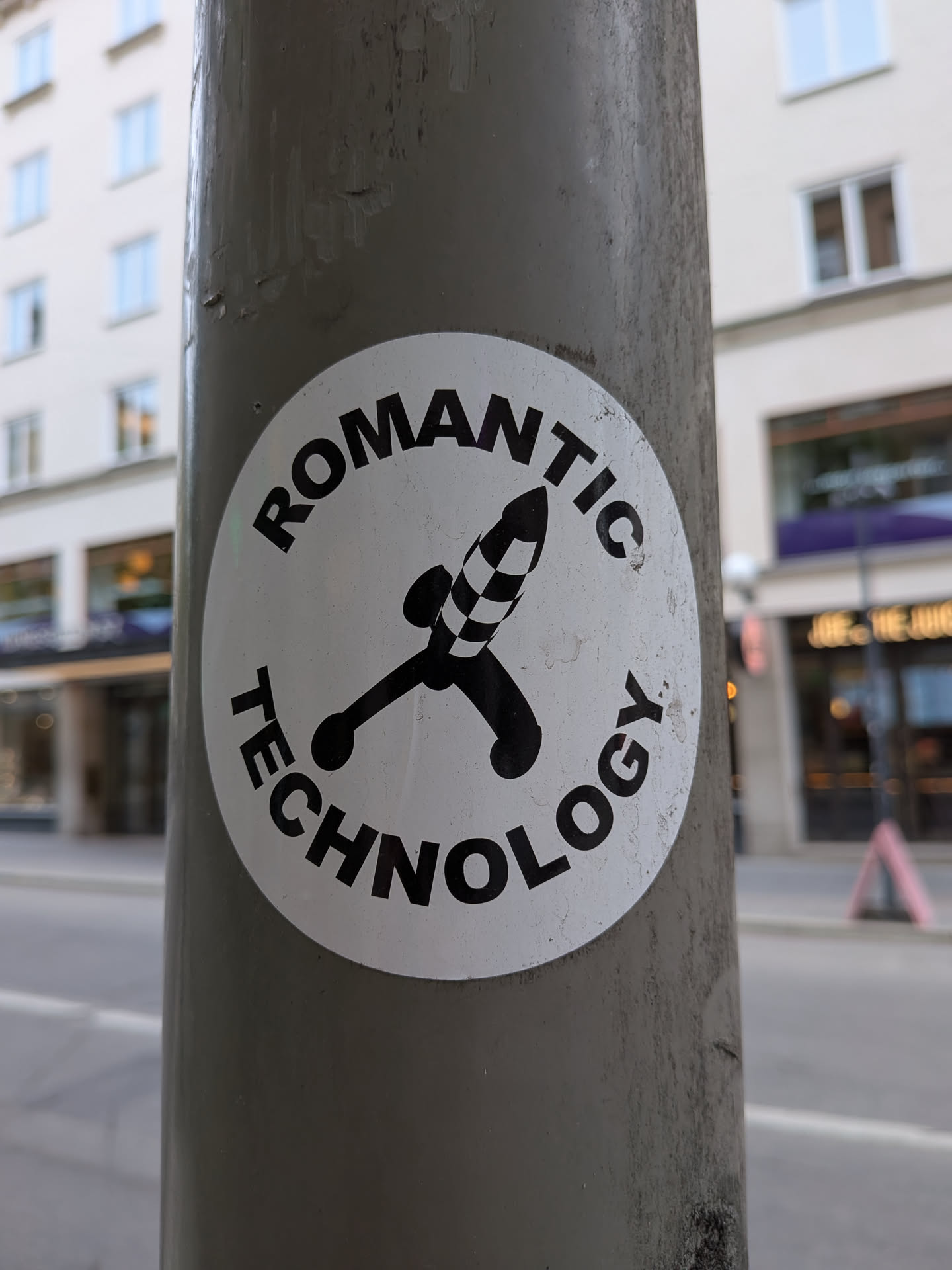 A circular white sticker featuring a bold black illustration of a stylized rocket or phallic object in the center. The object has a checkered pattern on its tip and two small spherical elements at its base, resembling a joystick or a dildo. Curved around the top and bottom edges of the circle, in black sans-serif capital letters, is the text 'ROMANTIC TECHNOLOGY'. The sticker shows signs of wear and dirt, indicating it's affixed to an outdoor pole.