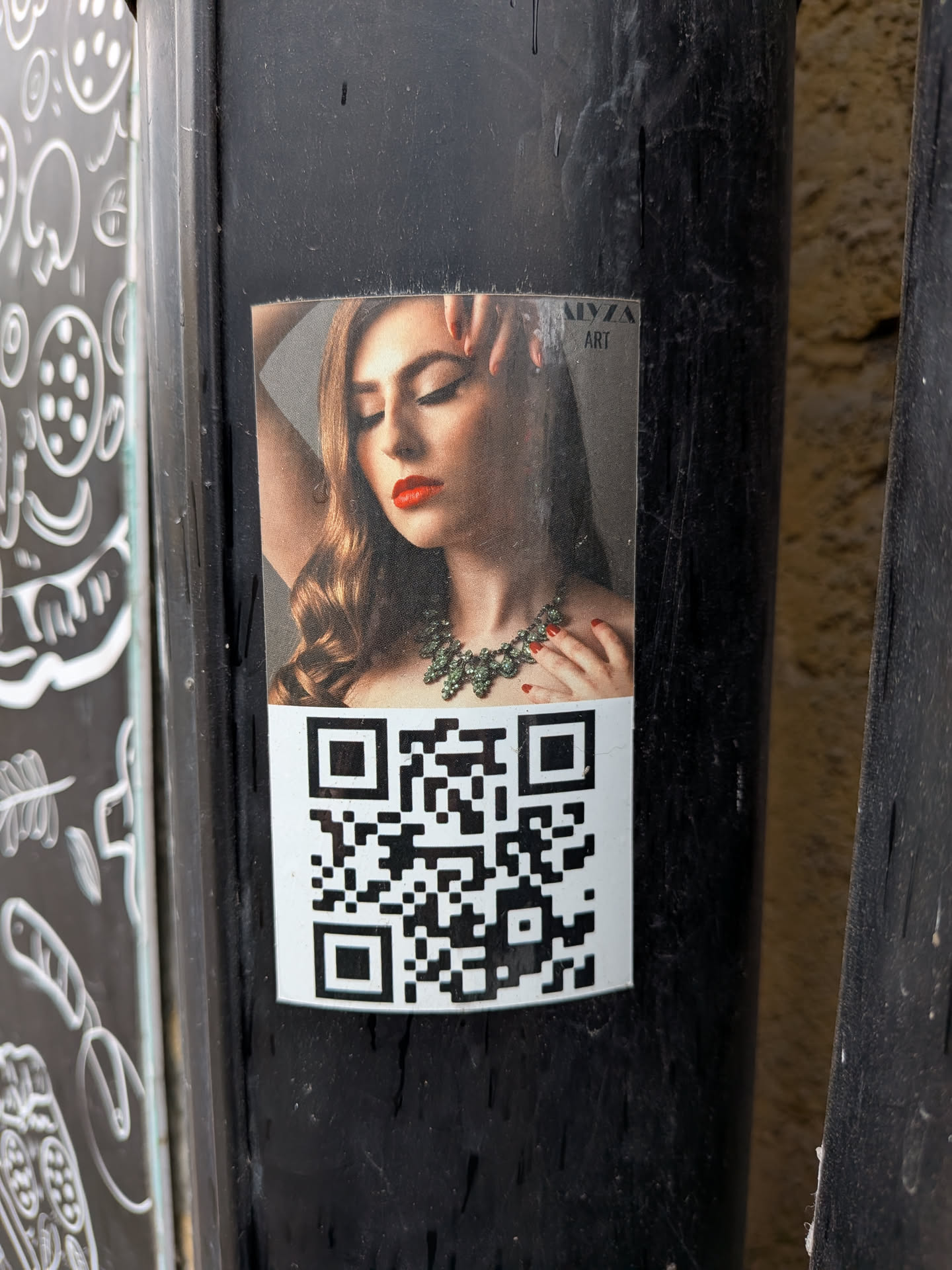 The sticker features a glossy, high-quality photograph of a woman with long, wavy brown hair, her eyes closed in a serene expression. She wears vibrant red lipstick and matching red nail polish on her visible hand, which is gently placed near her face. A prominent, intricate necklace made of green gemstones adorns her neck. The background of the portrait is dark and subtly textured. Below the portrait, a standard black and white QR code is printed on a white background, occupying the lower half of the sticker. The entire sticker is affixed to a dark, textured pole, showing some minor wear and tear around its edges.