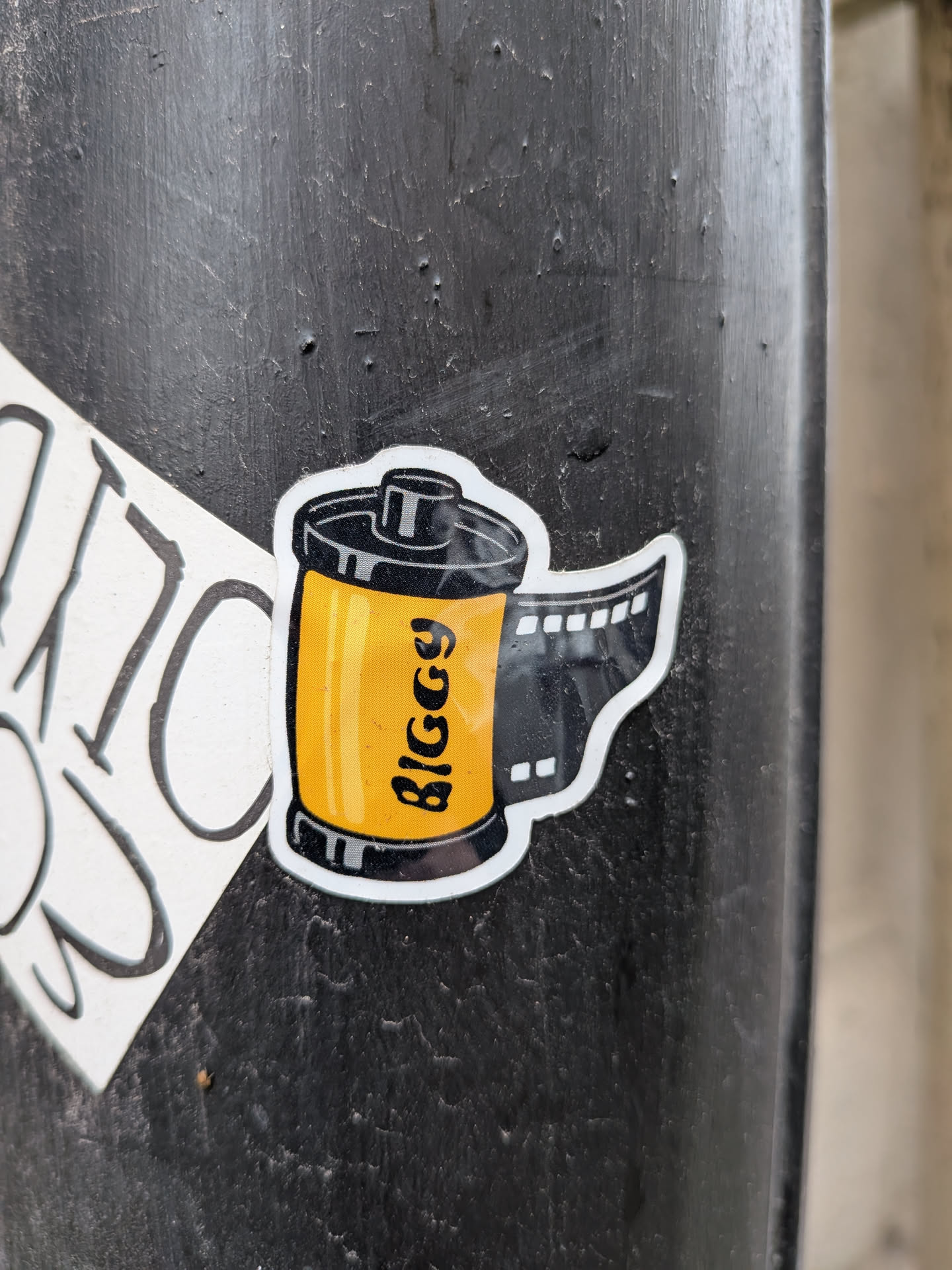 The sticker is a stylized illustration of a 35mm film roll. The main body of the roll is bright yellow with a textured pattern, and it has black rims and a black cap. Black text reading 'BIGGY' is vertically oriented on the yellow section. A strip of black film, complete with sprocket holes, extends from the side of the roll. The entire illustration is outlined in white.