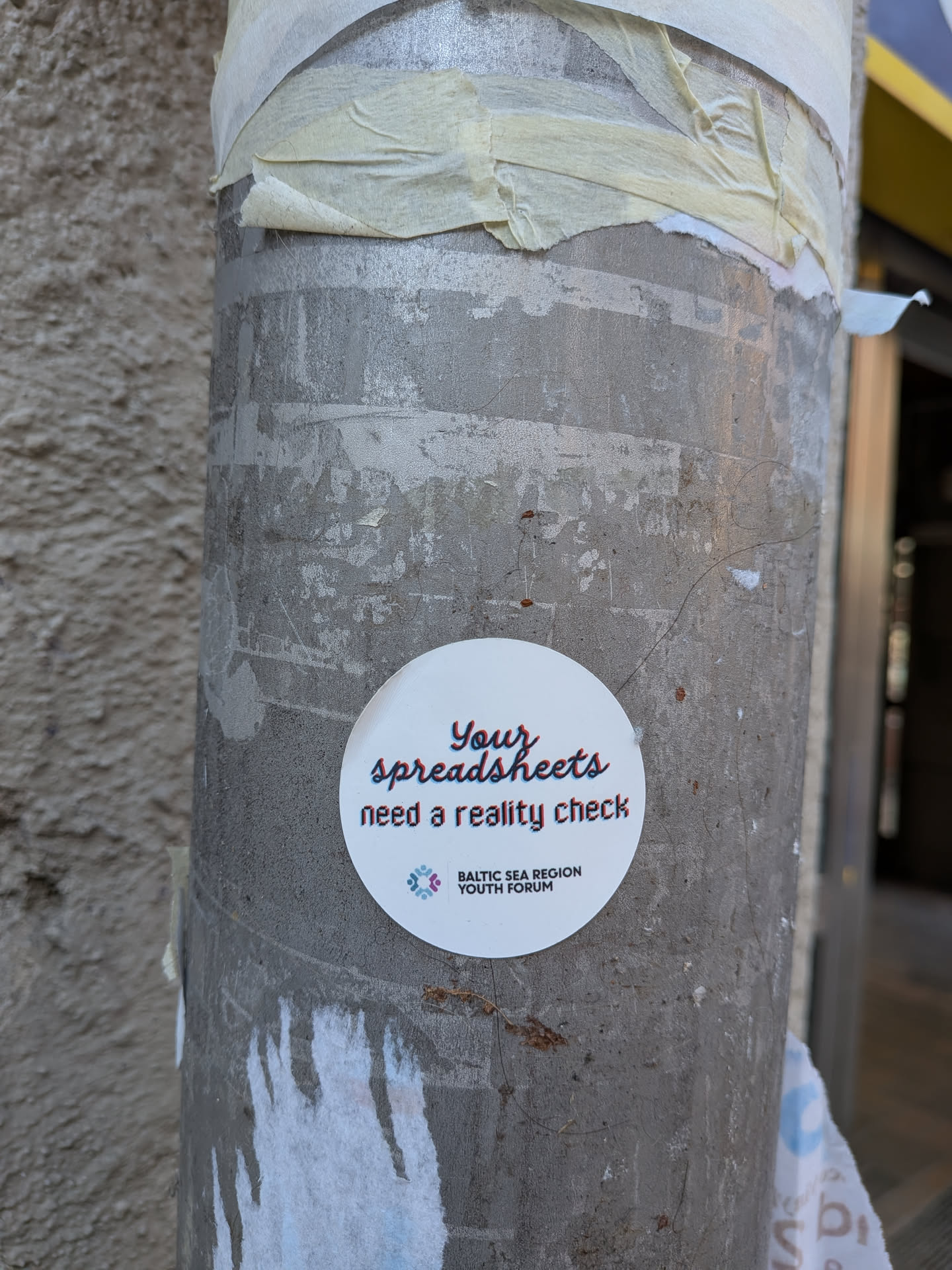 A circular white sticker featuring the phrase "Your spreadsheets need a reality check" in a stylized font with a red and blue glitch effect, giving it a slightly offset, 3D appearance. The word "Your" is predominantly red, "spreadsheets" is blue, and "need a reality check" is black with red and blue offsets. Below this text, there is a small logo depicting two stylized figures (one purple, one blue) connected in a circular pattern. Further below, in a smaller, black sans-serif font, it reads "BALTIC SEA REGION YOUTH FORUM". The sticker has a clean, modern design with a playful yet thought-provoking message.