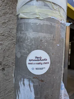 Street sticker A circular white sticker featuring the phrase "Your spreadsheets need a reality check" in a stylized font with a red and blue glitch effect, giving it a slightly offset, 3D appearance. The word "Your" is predominantly red, "spreadsheets" is blue, and "need a reality check" is black with red and blue offsets. Below this text, there is a small logo depicting two stylized figures (one purple, one blue) connected in a circular pattern. Further below, in a smaller, black sans-serif font, it reads "BALTIC SEA REGION YOUTH FORUM". The sticker has a clean, modern design with a playful yet thought-provoking message.