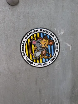 Street sticker A circular sticker featuring two cartoon animals, a grey mouse and a brown monkey-like creature, embracing warmly. The mouse has pink ears and tail, black eyes, and a white belly. The monkey-like character has a tan face, ears, and belly, with a wide, happy smile and brown fur. They are set against a background of vertical stripes, which are yellow and black on the left side, and blue and black on the right side. The central image is framed by a white ring containing black text. The top arc reads 'GAMMAL KÄRLEK ROSTAR ALDRIG', and the bottom arc reads 'ANTI 1891', flanked by small black leaf motifs. The entire design has a black outline and a white outer border, giving it a classic cartoon aesthetic.