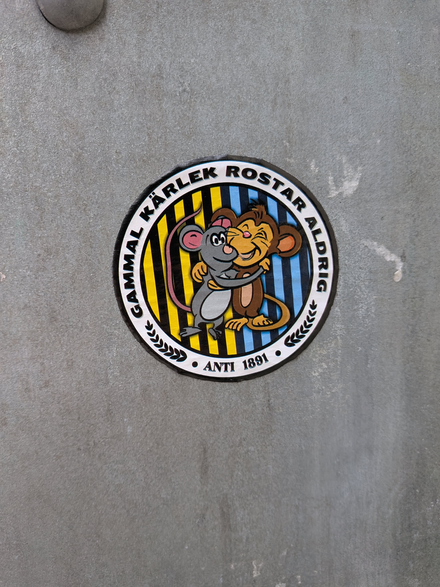A circular sticker featuring two cartoon animals, a grey mouse and a brown monkey-like creature, embracing warmly. The mouse has pink ears and tail, black eyes, and a white belly. The monkey-like character has a tan face, ears, and belly, with a wide, happy smile and brown fur. They are set against a background of vertical stripes, which are yellow and black on the left side, and blue and black on the right side. The central image is framed by a white ring containing black text. The top arc reads 'GAMMAL KÄRLEK ROSTAR ALDRIG', and the bottom arc reads 'ANTI 1891', flanked by small black leaf motifs. The entire design has a black outline and a white outer border, giving it a classic cartoon aesthetic.