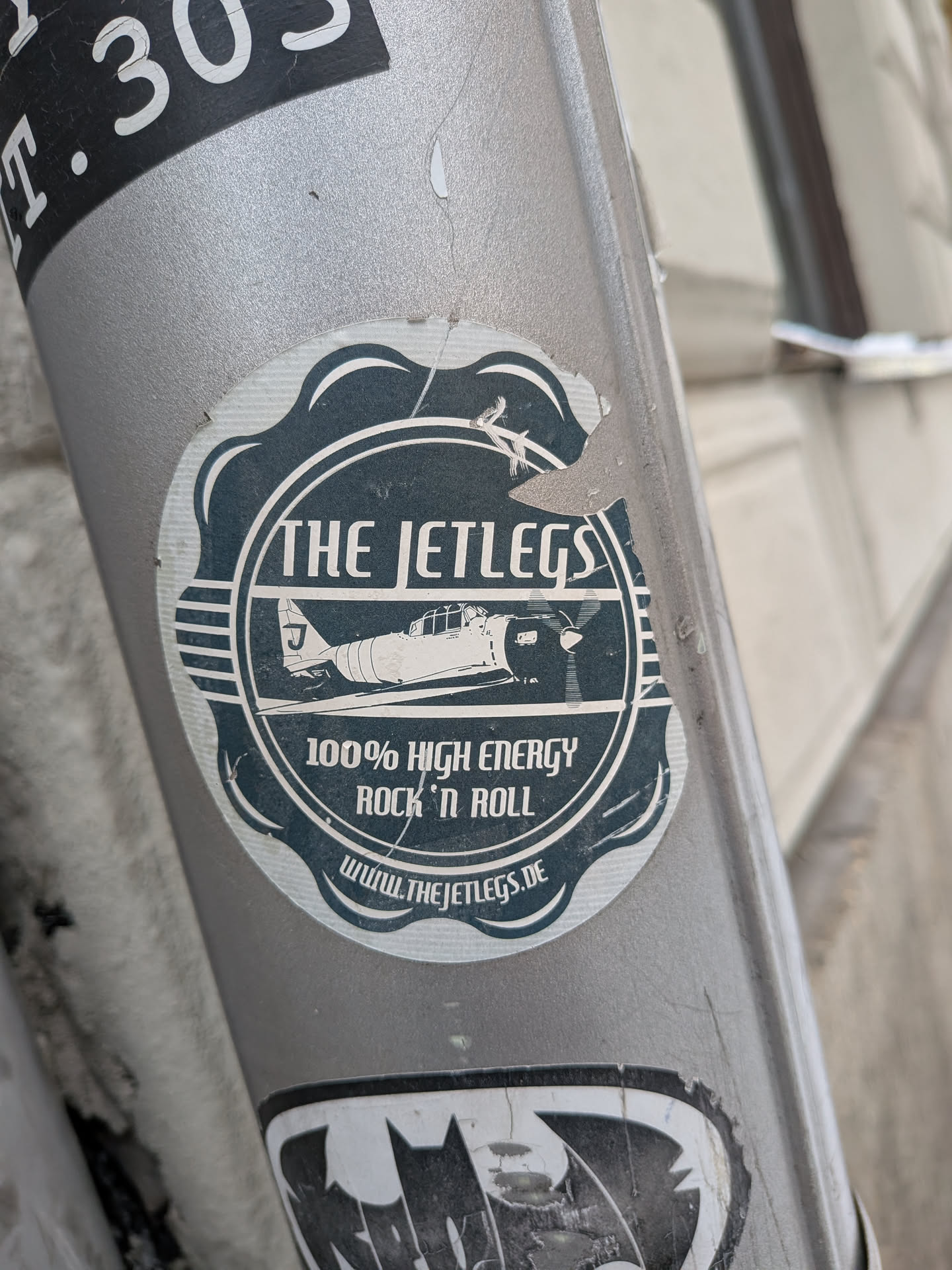 Street sticker The sticker is predominantly dark teal/blue and white, with a distressed, vintage aesthetic. It has a roughly circular shape with a decorative, scalloped outer edge. The central design features a stylized, vintage-era single-engine propeller airplane, depicted in white, flying horizontally. The letter 'J' is visible on the airplane's tail fin. Above the airplane, in bold white capital letters, is the band name "THE JETLEGS". Below the airplane, also in white capital letters, it reads "100% HIGH ENERGY ROCK 'N ROLL". At the very bottom, in smaller white text, is the website "WWW.THEJETLEGS.DE". The entire design is framed within a double-line white circle, and there are horizontal white stripes on either side of the airplane within this frame. The sticker shows signs of wear, with small tears and scuffs, particularly around the edges and top.