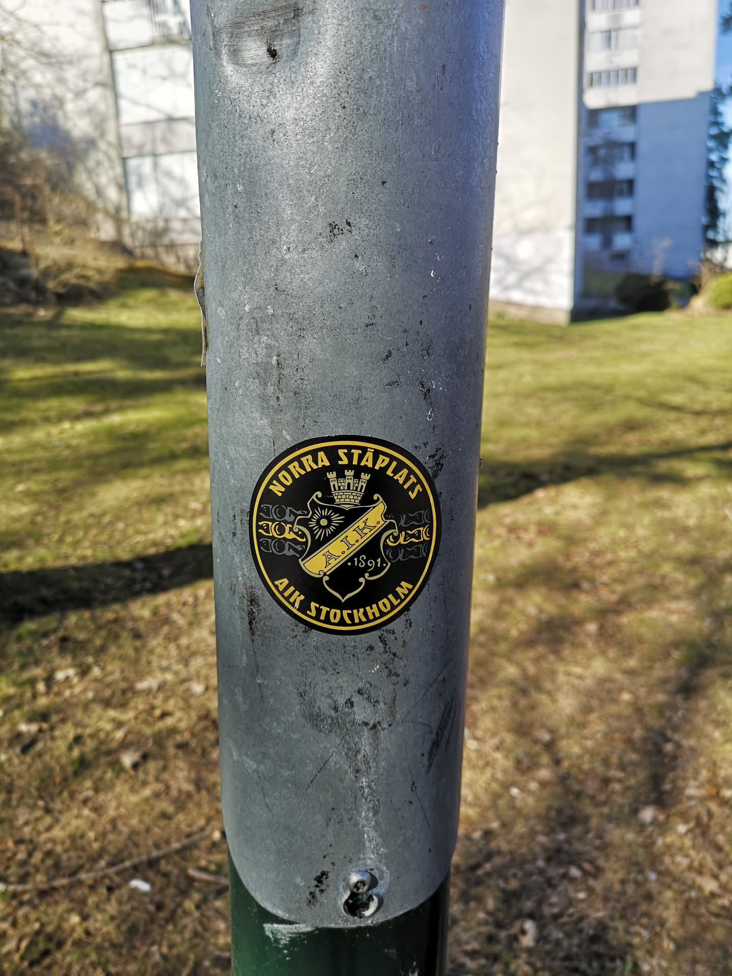 A circular sticker featuring the crest of AIK Stockholm, rendered in a striking black and gold/yellow color scheme. The outer edge of the sticker is a gold ring with black text. The central area is black, showcasing a gold shield-like emblem. At the top of this emblem is a gold crown with three turrets. Below the crown, a banner-like element contains the letters 'A.I.K.' in gold, with the year '1891' positioned directly beneath it. Ornate gold patterns flank the central shield. The top arc of the sticker reads 'NORRA STÅPLATS' in gold capital letters, and the bottom arc reads 'AIK STOCKHOLM', also in gold capital letters.