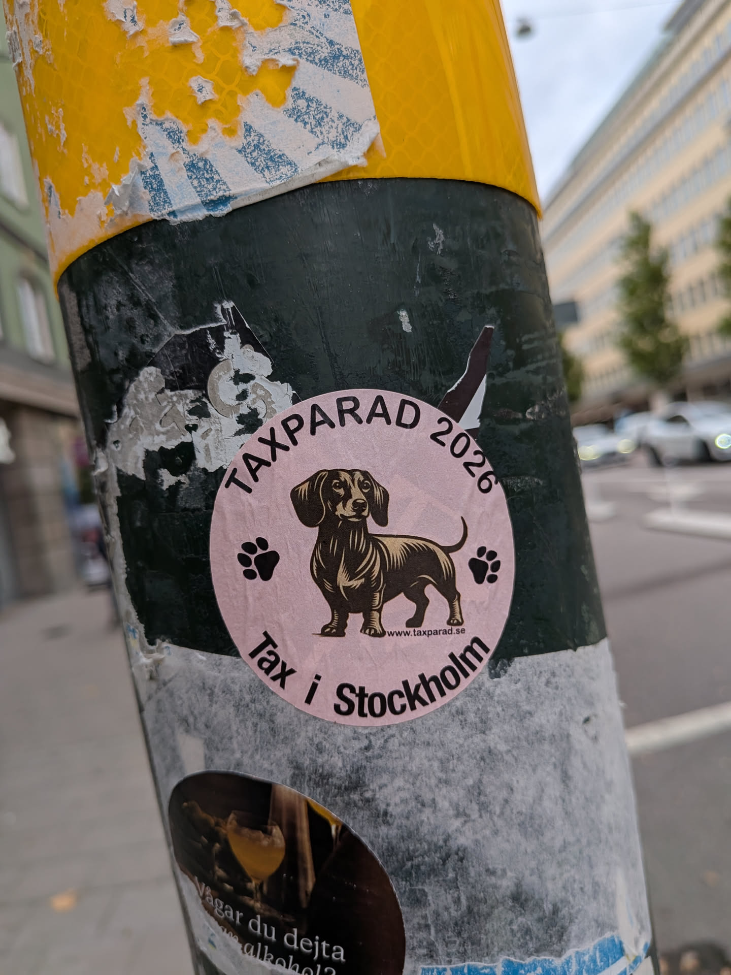 A circular, light pink sticker affixed to a dark green pole. The sticker features a detailed black and brown illustration of a dachshund standing in the center, flanked by two small black paw prints. The text 'TAXPARAD 2026' is printed in black along the top curve, and 'Tax i Stockholm' is printed along the bottom curve. A small website address, 'www.taxparad.se', is visible in tiny text near the dog's hind leg.