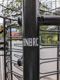 Street sticker A small rectangular sticker with a black background and bold, white, condensed sans-serif letters spelling 'BNBRC', affixed to a black metal post.