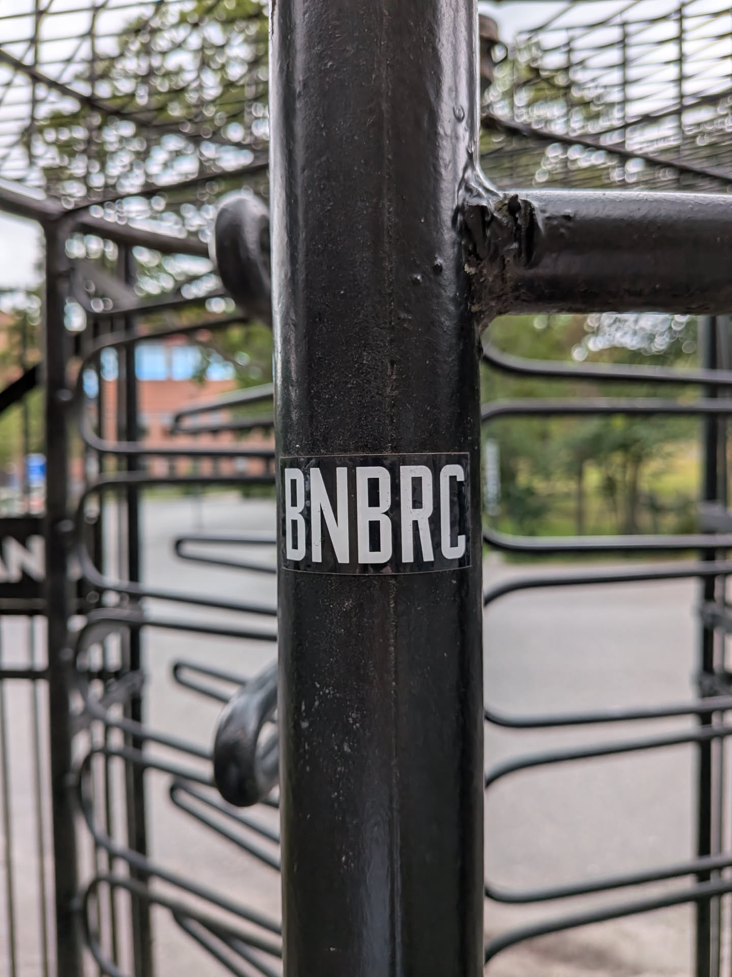 A small rectangular sticker with a black background and bold, white, condensed sans-serif letters spelling 'BNBRC', affixed to a black metal post.