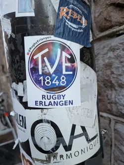 Street sticker The central sticker is a white, rectangular adhesive featuring a prominent circular logo. The logo's background is a vibrant galaxy scene, showcasing swirling purples, blues, and pinks, dotted with stars and a visible planet or moon. Overlaid on this cosmic backdrop are the bold white letters 'TVE' and the year '1848' below them. Above the circular logo, a small Instagram icon precedes the handle '@RugbyErlangen'. Below the circle, in a dark blue/purple font, are the words 'RUGBY' and 'ERLANGEN' stacked. The sticker appears slightly worn at the edges, indicating it's a physical sticker applied to a surface.