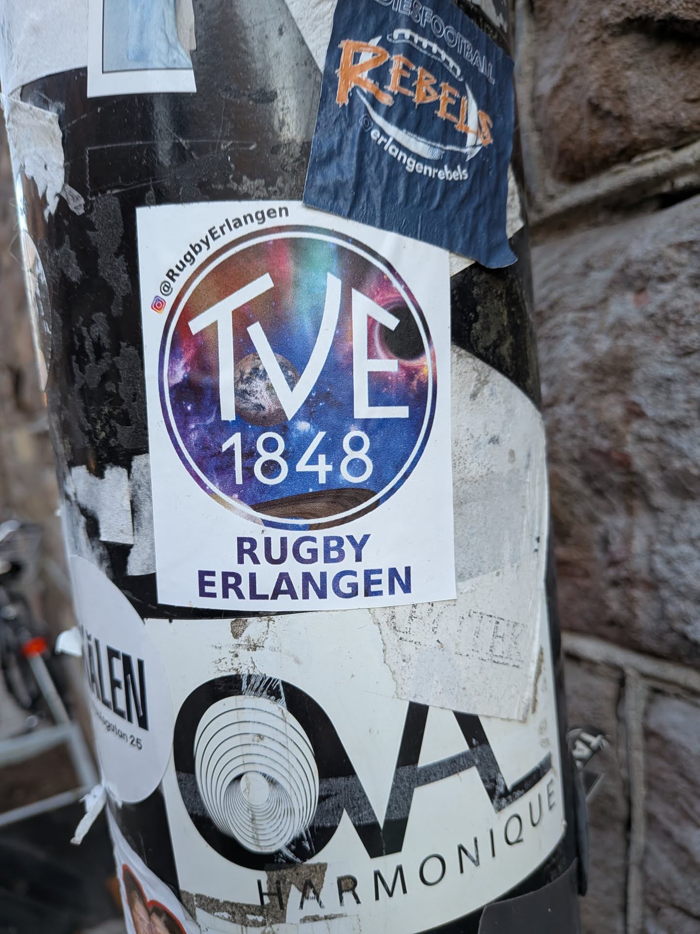 The central sticker is a white, rectangular adhesive featuring a prominent circular logo. The logo's background is a vibrant galaxy scene, showcasing swirling purples, blues, and pinks, dotted with stars and a visible planet or moon. Overlaid on this cosmic backdrop are the bold white letters 'TVE' and the year '1848' below them. Above the circular logo, a small Instagram icon precedes the handle '@RugbyErlangen'. Below the circle, in a dark blue/purple font, are the words 'RUGBY' and 'ERLANGEN' stacked. The sticker appears slightly worn at the edges, indicating it's a physical sticker applied to a surface.