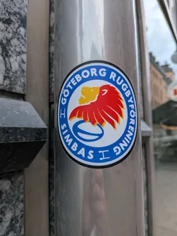 Street sticker Circular sports sticker with a blue outer ring containing white text. The center features a stylized yellow and red lion head above a blue rugby ball on a white background.