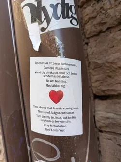 The sticker is rectangular with a white background. It features text in both Swedish and English. At the bottom center of the sticker is a prominent, bright red heart symbol. The text discusses the imminent coming of Jesus and the Day of Judgment, urging the reader to turn to Jesus for forgiveness of sins and pray for salvation. The sticker appears to be applied to a brown, cylindrical surface.