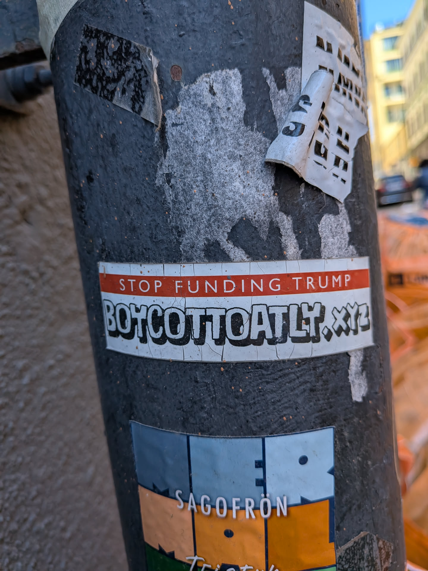 The sticker is a rectangular, horizontally oriented design with a white background. At the top, there's a prominent red stripe containing the text "STOP FUNDING TRUMP" in white, sans-serif capital letters. Below this, the main body of the sticker features the text "BOYCOTTOATLT.XYZ" in large, bold, black capital letters with a white outline, giving it a slightly three-dimensional appearance. The sticker shows signs of wear, with visible cracks and peeling, particularly on the white background and the black lettering, indicating it has been exposed to the elements. It is affixed to a dark, textured pole, with other faded stickers partially visible around it.