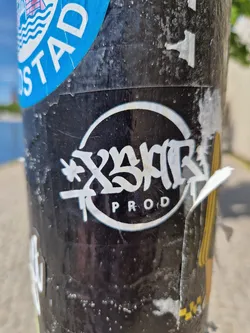 Street sticker A black and white circular sticker with stylized graffiti text that says "X3MR Prod." The style is reminiscent of street art and is applied to a weathered surface. 