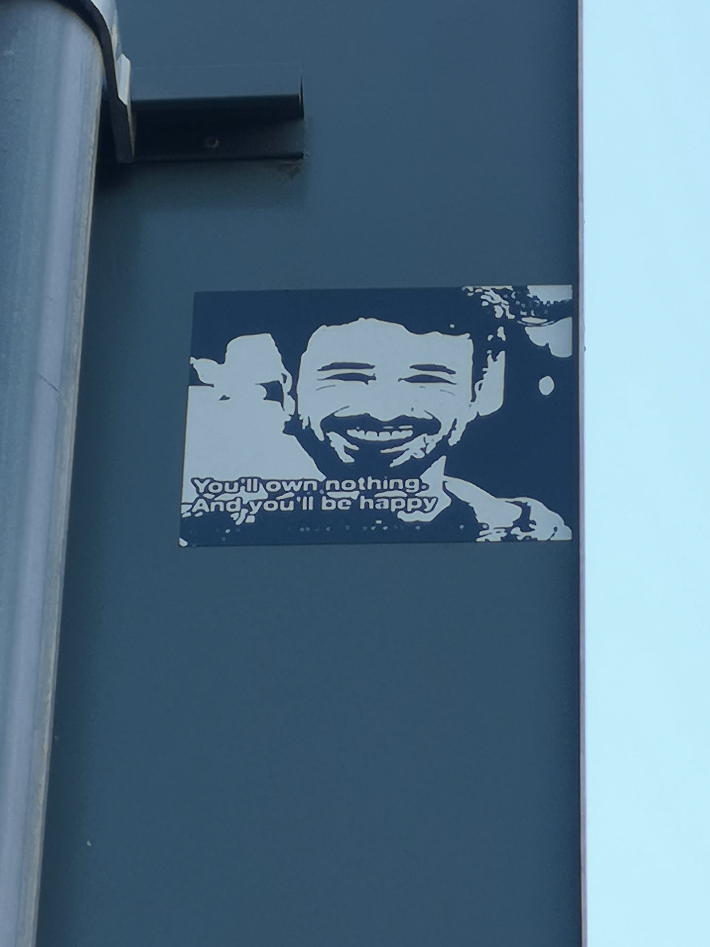 The sticker is a rectangular, two-tone graphic, predominantly dark blue/grey with light grey/white elements. It features a prominent, smiling man with short, curly hair and a beard, depicted in a high-contrast, stencil-like style. His eyes are closed in a wide smile, revealing teeth. Behind him, to the left, a less distinct, smiling silhouette of another person is visible. Below the main figure, the phrase "You'll own nothing. And you'll be happy." is printed in the same light grey/white color. The sticker is affixed to a dark blue-grey wall.