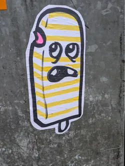 Street sticker This sticker features a whimsical illustration of a popsicle with yellow and white stripes. The popsicle has a cartoonish face with big eyes and a slightly frowning mouth. It has a small pink detail near the top. The style is simple and playful.