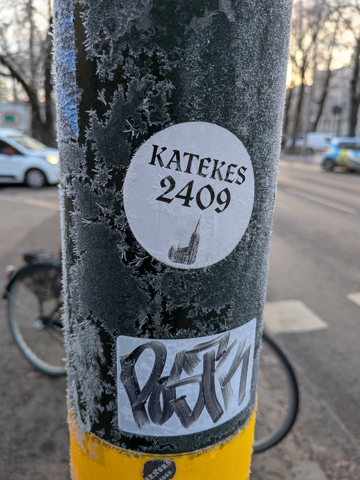 The central sticker is circular and white, with black text and an illustration. The text reads 'KATEKES' in a serif font at the top, and '2409' below it in a larger, bolder font. Beneath the numbers is a silhouette of a gothic-style building, possibly a church or cathedral, with a tall spire. The sticker appears slightly worn and is partially covered in frost. Below this circular sticker is another, larger sticker with black graffiti-style lettering on a white background. This graffiti is abstract and appears to be a tag.