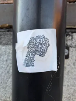 Street sticker The sticker is white and roughly rectangular with a slightly crumpled corner. At its center, a silhouette is depicted using dense, overlapping black lines. The lines vary in direction, creating a textured, abstract pattern within the outlined shape. The overall impression is minimalist and graphic.