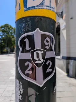 Street sticker The sticker is shaped like a classic shield or escutcheon, with a prominent white outline. The interior of the shield is divided into four quadrants by a light grey cross. The top-left quadrant contains the number '1', the top-right contains '9', the bottom-left contains '2', and the bottom-right contains '2'. These numbers are in a light grey color against a black background in their respective quadrants. In the very center of the grey cross, there is a smaller, darker grey shield with a white outline, containing a stylized white letter that resembles a gothic 'G' or 'Q'. The overall color scheme is black, various shades of grey, and white, giving it a clean, emblem-like appearance. The sticker has a slight reflective sheen.
