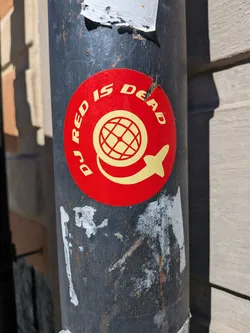 Street sticker A circular sticker with a vibrant red background features yellow (or gold) text and imagery. The text 'DJ RED IS DEAD' is arranged in an arc around the top half of the circle. In the center, there is a stylized globe icon with grid lines, and beneath it, a yellow silhouette resembling an airplane or rocket appears to be circling the lower part of the globe. The sticker shows some signs of wear, with minor tears and scuffs, and is affixed to a dark, weathered pole.