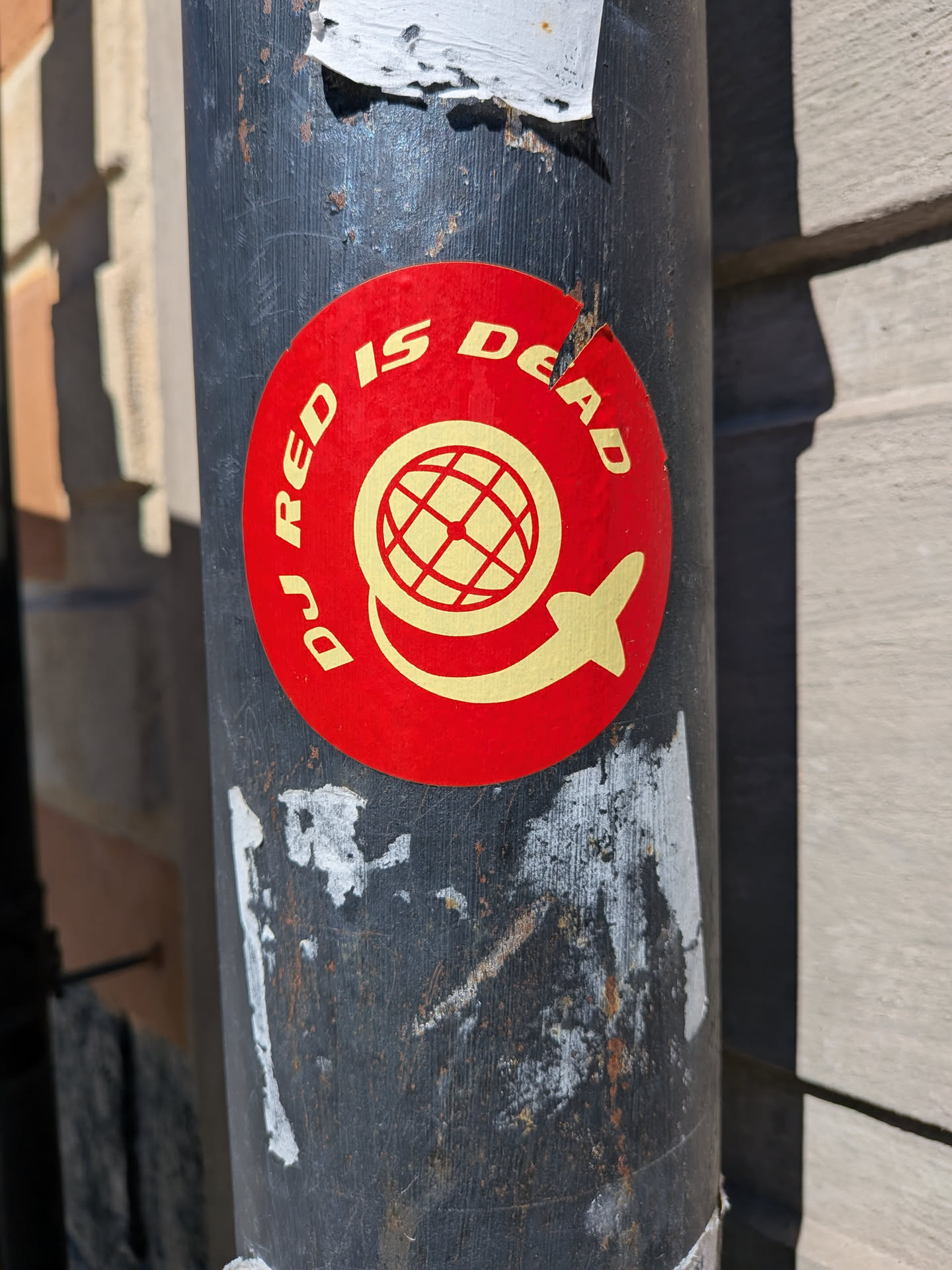 A circular sticker with a vibrant red background features yellow (or gold) text and imagery. The text 'DJ RED IS DEAD' is arranged in an arc around the top half of the circle. In the center, there is a stylized globe icon with grid lines, and beneath it, a yellow silhouette resembling an airplane or rocket appears to be circling the lower part of the globe. The sticker shows some signs of wear, with minor tears and scuffs, and is affixed to a dark, weathered pole.