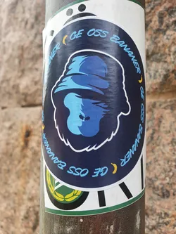 Street sticker A round sticker with a blue monkey wearing a bucket hat. The text around the monkey says "Ge oss bananer" which translates to "Give us bananas" in Swedish. 