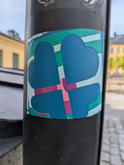 A square sticker featuring a stylized blue four-leaf clover silhouette. The design is intersected by geometric stripes in green, pink, and light blue, creating a colorful mosaic effect.