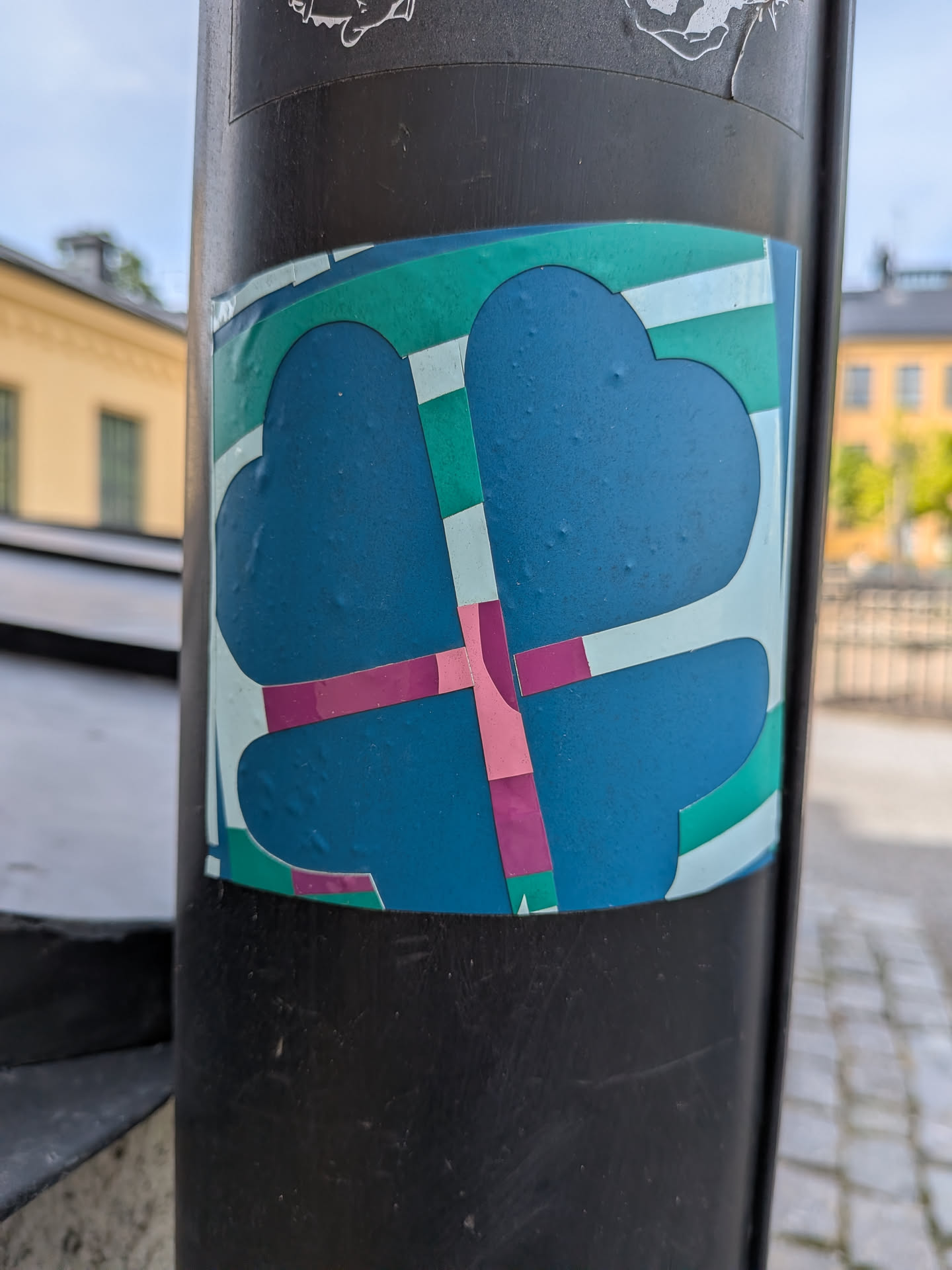 A square sticker featuring a stylized blue four-leaf clover silhouette. The design is intersected by geometric stripes in green, pink, and light blue, creating a colorful mosaic effect.