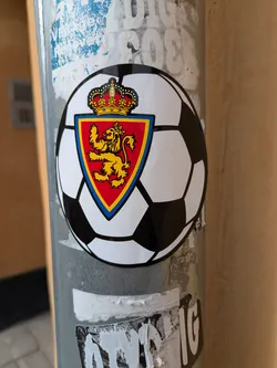A round sticker attached to a pole, features a soccer ball design. In the center, there is a red shield bordered with blue. Inside the shield, a golden lion stands prominently, and a golden crown sits atop the shield. The design suggests a soccer club or organization with royal connections.