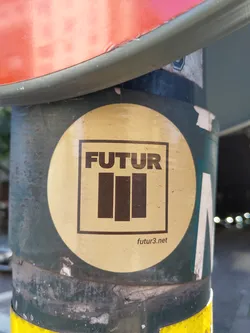 Street sticker Round beige sticker with black text and logo. The text 'FUTUR' is written in uppercase letters inside a square, followed by three vertical bars below it. At the bottom, a website address 'futur3.net' is printed in a smaller font. The sticker is applied to a dark green pole.