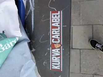 Street sticker A vertically oriented rectangular sticker featuring bold white block letters with black outlines against a red background, framed by a thin green and white border. The text reads 'KURVA CARLABEL', with a stylized cartoon face wearing a red beanie positioned between the two words. The face has simple features: small black dot eyes, a short line for a nose, and a slightly curved line for a mouth, all outlined in black. The letters have small square cutouts, adding to their unique style.