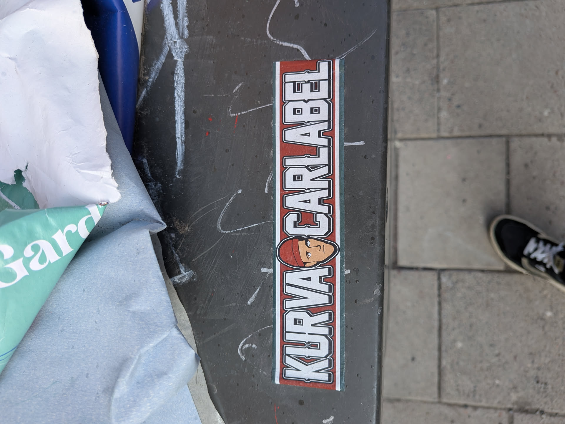 A vertically oriented rectangular sticker featuring bold white block letters with black outlines against a red background, framed by a thin green and white border. The text reads 'KURVA CARLABEL', with a stylized cartoon face wearing a red beanie positioned between the two words. The face has simple features: small black dot eyes, a short line for a nose, and a slightly curved line for a mouth, all outlined in black. The letters have small square cutouts, adding to their unique style.