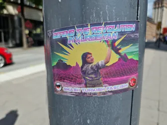 Street sticker A sticker featuring a person raising their fist in front of a stylized landscape. The text on the sticker says "DEFEND THE REVOLUTION IN KURDISTAN" and "SMASH TURKISH FASCISM".