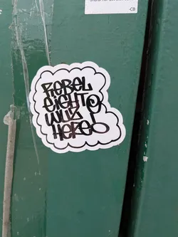 Street sticker A die-cut, cloud-shaped sticker with a white background and a thin black border. The center features stylized, hand-written graffiti text in black ink that reads 'REBEL EIGHT WUZ HERE!'. The sticker is affixed to a textured, green-painted surface.