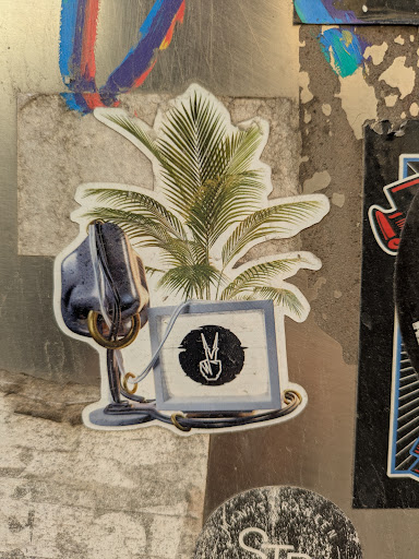 The sticker shows a vintage microphone next to a palm tree, below of which is a screen showing a symbol of a peace sign. This symbol appears to be in a circular frame. The elements are connected by metallic-looking rings and wires.