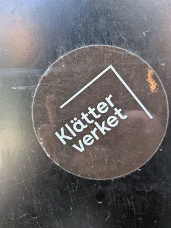 Street sticker A circular sticker with a dark brown, slightly reflective background. The text "Klätter verket" is prominently displayed in white, sans-serif font, angled slightly upwards from left to right. Above and to the right of the text, there is a white, thick, angular line forming an open corner, resembling an incomplete square or a stylized 'L' shape. The sticker shows signs of wear, including scratches and some dirt, indicating it's been applied for some time on a dark, reflective surface.