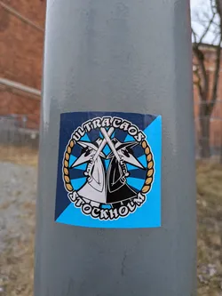 Street sticker A circular sticker featuring two stylized figures, reminiscent of the 'Spy vs. Spy' characters, one in white and one in black, mirroring each other. Both figures wear pointed hats and dark sunglasses, and each holds a white baseball bat crossed in front. The background within the circle is light blue with dark blue star-like or triangular accents. A golden braided rope forms a border around the central figures. Arched text above reads 'ULTRACHAOS' and below reads 'STOCKHOLM', both in white with a black outline. The circular design is set against a larger square background that is diagonally split between dark blue and light blue. The sticker is applied to a grey pole.
