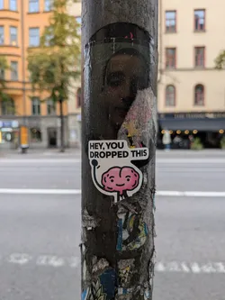 Street sticker A die-cut sticker on a pole depicting a smiling pink cartoon brain waving, with a speech bubble reading 'HEY, YOU DROPPED THIS'.