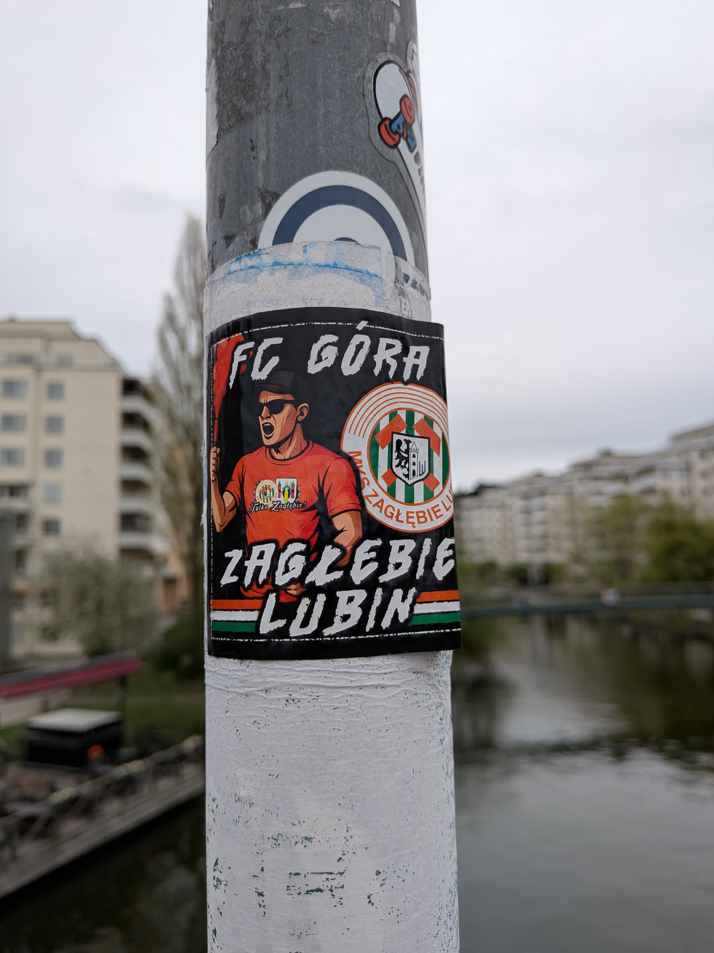 A rectangular sticker featuring a stylized illustration of a male football fan from the chest up, set against a black background. The fan wears sunglasses and an orange t-shirt, with an open-mouthed, shouting expression. He holds a red flare or pyrotechnic device in his right hand, emitting red smoke. On his t-shirt, a small badge reads 'Tylko Zagłębie!'. To the right of the fan is a circular football club crest with green and white vertical stripes, an orange border, and a black shield containing a building (possibly a castle or tower) and a tree. The text 'MKS ZAGŁĘBIE LUBIN' is partially visible around the crest. Above the fan, in distressed white font, is 'FC GÓRA'. Below the fan and crest, also in distressed white font, is 'ZAGŁĘBIE LUBIN'. The bottom edge of the sticker features a horizontal stripe pattern in orange, white, and green.