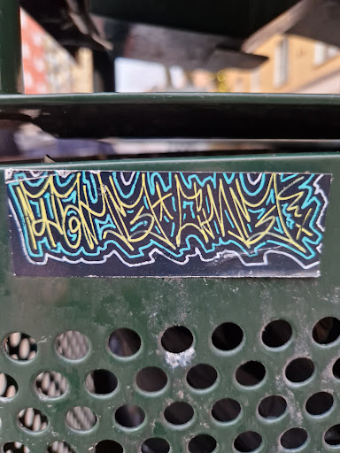 Street sticker A sticker with an abstract graffiti design in yellow, light blue and teal on a black background is affixed to a dark-green perforated metal surface. The graffiti-style design is predominantly composed of stylized lines and curves.