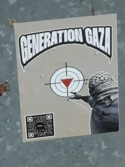 Street sticker A rectangular sticker with a light grey background featuring a central graphic. At the top, the words "GENERATION GAZA" are displayed in large, bold, white letters with a thick black outline, forming an arch. Below this, a target symbol with concentric white and black circles and crosshairs is prominent, with a solid red inverted triangle in the bullseye. To the right, a grayscale figure, from the chest up, is depicted wearing a keffiyeh that covers their face, holding a slingshot and aiming towards the target. In the bottom left corner, there is a black and white QR code with the letters "GNG" visible in its center.
