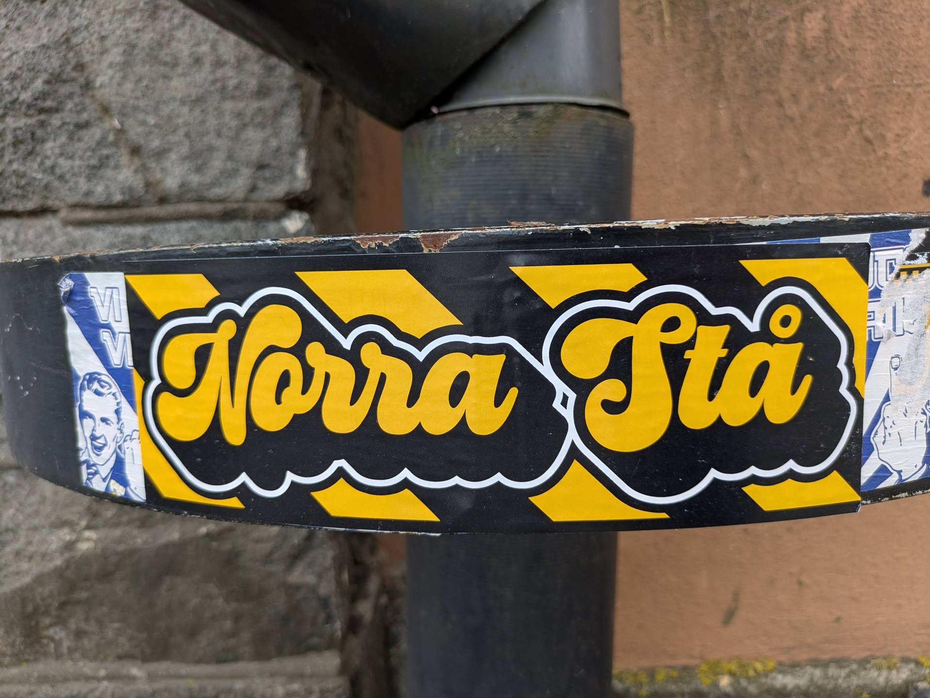 Street sticker The sticker is horizontally oriented, featuring prominent yellow text "Norra Stå" with a thick white outline, set against a black background with diagonal yellow stripes, reminiscent of caution tape. The text itself is in a rounded, bubbly, script-like font. The sticker is applied to a dark, possibly metal, pipe or drainpipe, showing some wear and slight peeling at the edges, indicating its physical presence. Remnants of other stickers are visible on either side, partially covered by this one; on the far left, a portion of a blue and white sticker with a smiling male face is visible, and on the far right, a faded white and blue sticker with a hand gesture is partially seen.