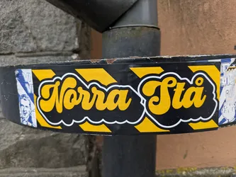 Street sticker The sticker is horizontally oriented, featuring prominent yellow text "Norra Stå" with a thick white outline, set against a black background with diagonal yellow stripes, reminiscent of caution tape. The text itself is in a rounded, bubbly, script-like font. The sticker is applied to a dark, possibly metal, pipe or drainpipe, showing some wear and slight peeling at the edges, indicating its physical presence. Remnants of other stickers are visible on either side, partially covered by this one; on the far left, a portion of a blue and white sticker with a smiling male face is visible, and on the far right, a faded white and blue sticker with a hand gesture is partially seen.