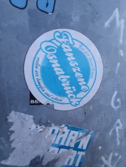 A round sticker with the text "Fanszene Osnabrück" in a stylized font.  The words "Nur für diesen Verein" (only for this club) and "wollen wir kämpfen und schrein" (we want to fight and scream) are also visible in smaller text. The sticker is light blue and white and is affixed to a gray surface.