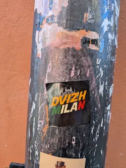 Street sticker A rectangular sticker with a black background. At the top, a white silhouette of the Milan Cathedral (Duomo di Milano) is prominently featured. Below the silhouette, the word 'DVIZH' is written in bold, orange-yellow capital letters. Underneath 'DVIZH', the word 'MILAN' is displayed in capital letters, with each letter colored to represent the Italian flag: 'M' in green, 'I' in a light cream/off-white, 'L' in a light cream/off-white, 'A' in a light cream/off-white, and 'N' in red. The sticker has slightly worn edges and is applied to a textured, peeling pole.