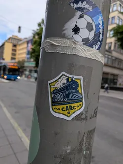 Street sticker A yellow and blue shield sticker depicting a vintage steam train blowing smoke. The bottom features the acronym 'CARC' and small cursive text on a yellow textured background.