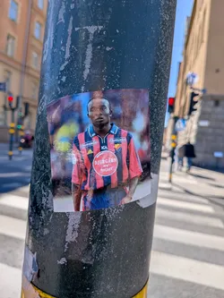 Street sticker The sticker features a portrait of a dark-skinned male football player with short, dark hair, looking directly forward. He is wearing a red and blue striped jersey with a blue collar and blue shorts. Prominently displayed on the jersey are sponsor logos: 'ICA' above the chest, 'Quesada' on the left shoulder, and a small Adidas logo on the right shoulder. A large red circular badge on the chest contains the text 'DJURGÅRDEN BY NIGHT' in white, along with the Djurgården IF crest. The background is a blurred mix of blue and reddish-brown, possibly indicating a stadium or field. The sticker is rectangular, appears to be a printed photograph, and shows some signs of wear and tear, with slight peeling at the edges, affixed to a dark, textured pole.