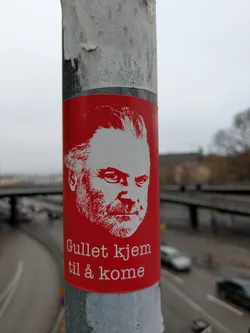 Street sticker A sticker with a red background on a pole. The sticker has a white line art drawing of a man's face at the top and the text "Gullet kjem til å kome" at the bottom.