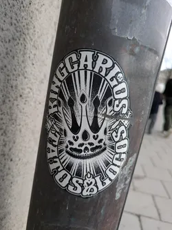 Street sticker A round black and white sticker adhered to a pole features the text "KING CARLOS" around the outer edge, surrounding a central design of a crown resembling a skull. The design is partially obscured by a rip in the sticker. Small skull icons are embedded around the edge of the design.